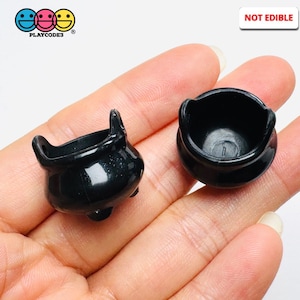 May include: Two small, black plastic cauldrons. One cauldron has a handle and three small feet. The other cauldron is round with a wide opening. The text "NOT EDIBLE" is in the upper right corner.