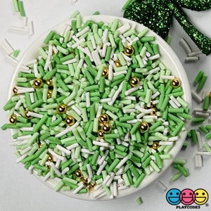 May include: A white ceramic bowl filled with green, light green, and white sprinkles, along with small gold-colored spherical beads. The sprinkles are in a cylindrical shape. The image is brightly lit and the bowl is on a white surface.
