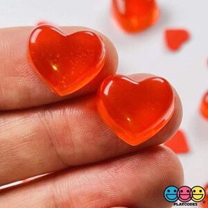 May include: Two bright orange heart-shaped candies, made of clear, translucent material, are held in a hand. The candies are slightly glossy and have a smooth surface.