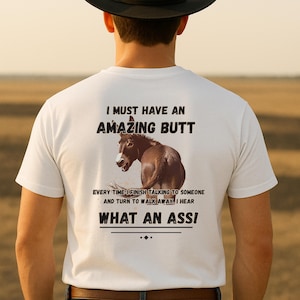May include: White t-shirt with the text "I MUST HAVE AN AMAZING BUTT" above a brown donkey illustration. Below the image, the text reads "EVERY TIME I FINISH TALKING TO SOMEONE AND TURN TO WALK AWAY, I HEAR WHAT AN ASS!"
