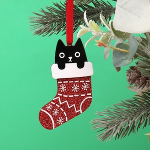 Può includere: Un ornamento a forma di calza di Natale rosso e bianco con un gatto nero che spunta dalla parte superiore. La calza ha una finitura scintillante e un bordo bianco.