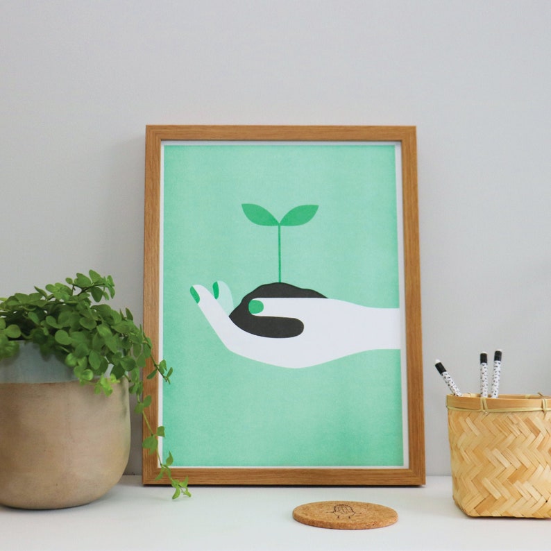Op de afbeelding: Een ingelijst print met een groene achtergrond waarop een gestileerde witte hand te zien is die een klein groen plantje vasthoudt dat uit zwarte aarde groeit.
