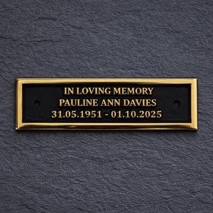 May include: A rectangular black plaque with a gold border, bearing the inscription "IN LOVING MEMORY" above "PAULINE ANN DAVIES" and the dates 31.05.1951 - 01.10.2025. The plaque is set against a dark grey, textured background.