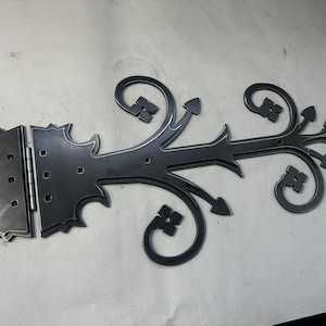 Peut inclure: Une charnière en métal noir avec un design décoratif. La charnière est composée de trois pièces : une pièce droite avec une extrémité pointue, une pièce incurvée avec un motif floral et une pièce rectangulaire avec une charnière.