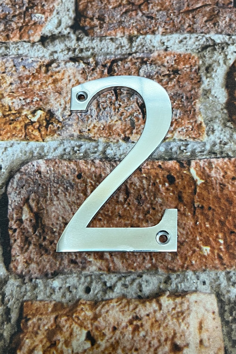 K&ouml;nnte beinhalten: Eine silberne Hausnummer zwei aus Metall. Die Nummer befindet sich auf einem Ziegelhintergrund.