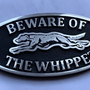 May include: A black and silver oval sign with the text "Beware of the Whippet" and a silver silhouette of a whippet running.