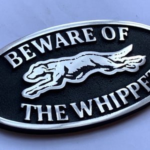 May include: Black and silver oval sign with the text "Beware of the Whippet" and a stylized image of a whippet dog running.