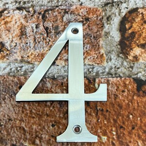 K&ouml;nnte beinhalten: Eine silberne Hausnummer vier aus Metall. Die Nummer ist an einer Backsteinwand befestigt.