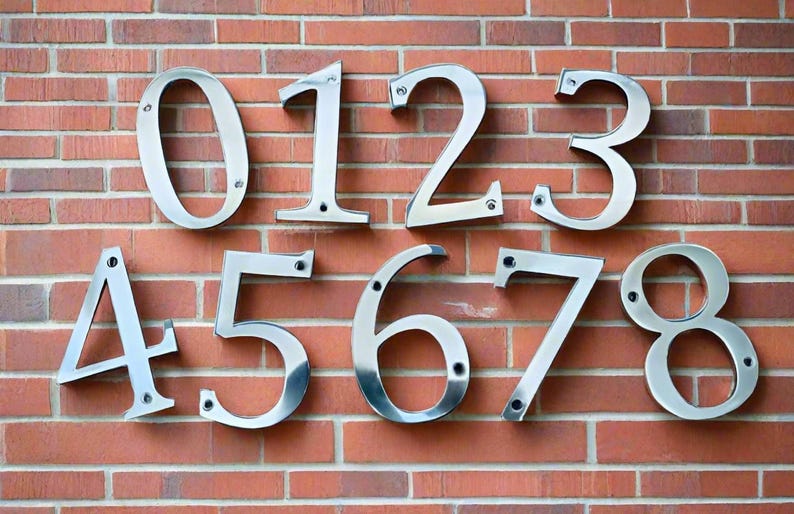 K&ouml;nnte beinhalten: Silberne Hausnummern 012345678 an einer Backsteinwand.
