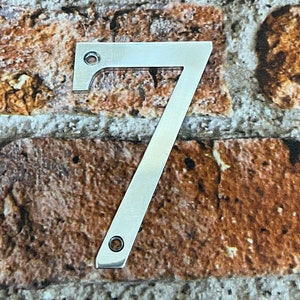 K&ouml;nnte beinhalten: Eine silberne Hausnummer sieben aus Metall. Die Nummer befindet sich vor einem Hintergrund aus Ziegelmauer.