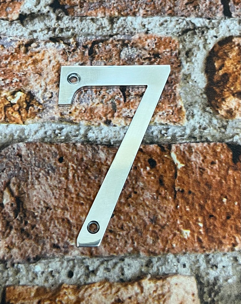 K&ouml;nnte beinhalten: Eine silberne Hausnummer sieben aus Metall. Die Nummer befindet sich vor einem Hintergrund aus Ziegelmauer.