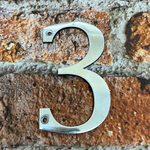 K&ouml;nnte beinhalten: Eine silberne Hausnummer drei aus Metall. Die Nummer ist an einer Backsteinwand befestigt.