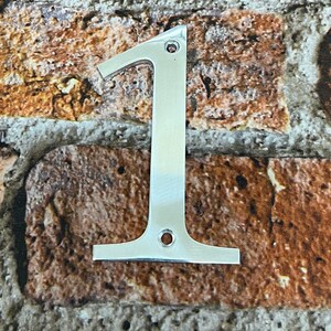 K&ouml;nnte beinhalten: Eine silberne Hausnummer eins aus Metall. Die Nummer ist an einer Backsteinwand befestigt.
