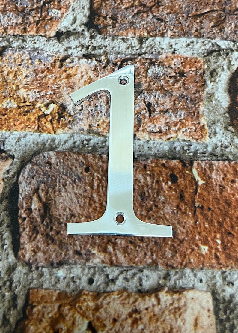 K&ouml;nnte beinhalten: Eine silberne Hausnummer eins aus Metall. Die Nummer ist an einer Backsteinwand befestigt.