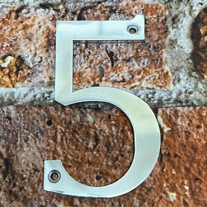 K&ouml;nnte beinhalten: Eine silberne Hausnummer f&uuml;nf aus Metall. Die Nummer ist an einer Backsteinwand befestigt.