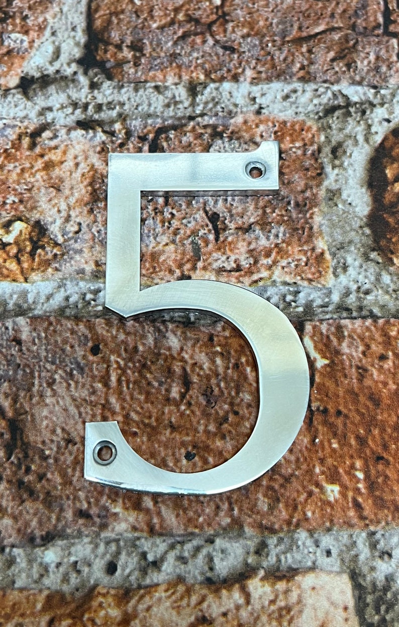 K&ouml;nnte beinhalten: Eine silberne Hausnummer f&uuml;nf aus Metall. Die Nummer ist an einer Backsteinwand befestigt.