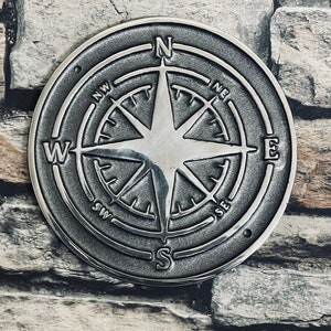 May include: A silver compass with a detailed design and raised lettering. The compass points north, south, east, and west. The compass is circular and has a raised star in the center.