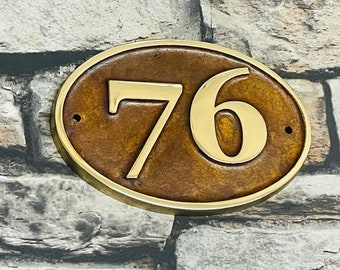 May include: Oval-shaped brass house number plaque with the number 76 in gold lettering on a brown background.