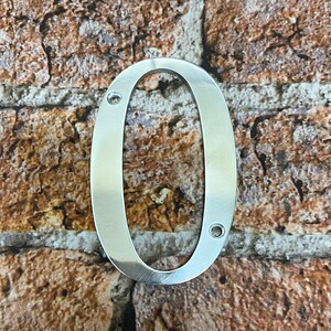 K&ouml;nnte beinhalten: Silberne Hausnummer Null aus Metall, mit zwei kleinen L&ouml;chern zur Befestigung. Die Nummer befindet sich vor einem Hintergrund aus Ziegelmauer.