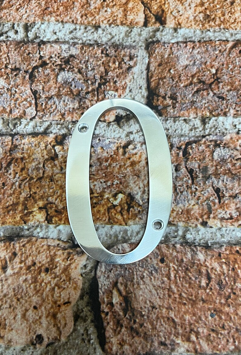 K&ouml;nnte beinhalten: Silberne Hausnummer Null aus Metall, mit zwei kleinen L&ouml;chern zur Befestigung. Die Nummer befindet sich vor einem Hintergrund aus Ziegelmauer.