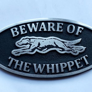 May include: A black oval metal sign with silver lettering that reads "Beware of the Whippet". A silver silhouette of a whippet dog is running across the sign.