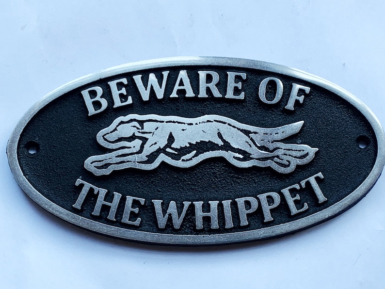 May include: A black oval metal sign with silver lettering that reads "Beware of the Whippet". A silver silhouette of a whippet dog is running across the sign.