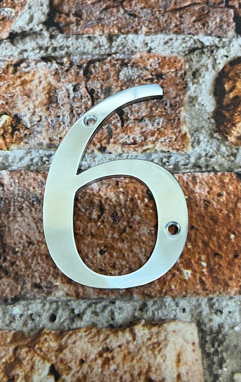 K&ouml;nnte beinhalten: Eine silberne Hausnummer sechs aus Metall. Die Nummer befindet sich auf einem braunen Ziegelhintergrund.
