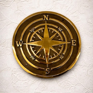 May include: A round, antique brass compass rose with directional markings (N, S, E, W, NE, NW, SE, SW). The compass is set against a textured white background. The compass rose is a decorative nautical element.