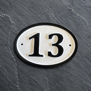 Op de afbeelding: Een ovaalvormig huisnummerbordje met het getal 13 in een vetgedrukt zwart lettertype op een witte achtergrond. Het bordje heeft een dikke zwarte rand en twee kleine ronde gaten aan de linker- en rechterkant voor montage.