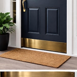 Puede incluir: Una puerta de entrada azul oscuro con una placa protectora de latón y un tirador. Una alfombra de fibra de coco marrón rectangular está colocada frente a la puerta. Una planta en maceta está a la izquierda de la puerta.