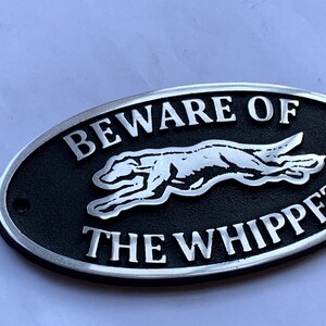May include: A black and silver oval-shaped metal sign with the text "Beware of the Whippet" and a silver silhouette of a whippet dog running.
