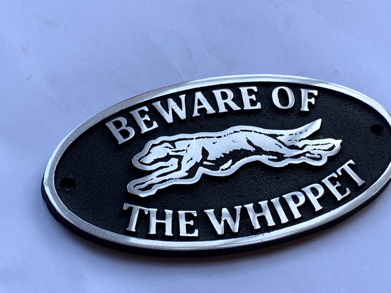 May include: A black and silver oval-shaped metal sign with the text "Beware of the Whippet" and a silver silhouette of a whippet dog running.