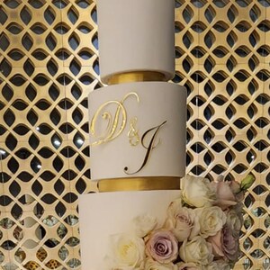 May include: A white wedding cake with four tiers decorated with a cascade of blush and ivory roses. The top tier has a gold ribbon and the initials "D & J" in gold script.