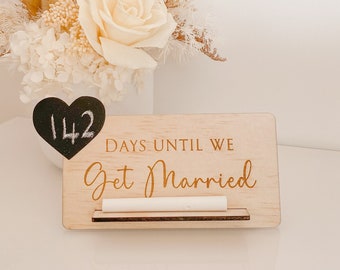 May include: A wooden sign with the text "Days Until We Get Married" and a chalkboard heart that says "142". The sign is sitting in front of a bouquet of dried flowers in a white vase.
