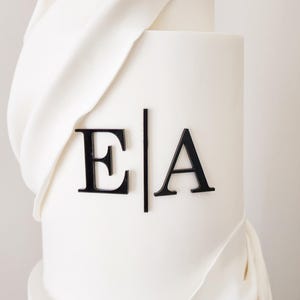 May include: A white wedding cake with a white fondant drape and black acrylic letters spelling "E|A" on the top tier.