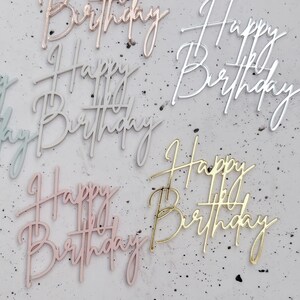 May include: Five acrylic cake toppers in different colours, each with the words "Happy Birthday" written in a cursive script. The colours are rose gold, silver, gold, light blue, and pink.