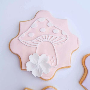 May include: A pink and white sugar cookie decorated with a white icing mushroom and a white flower. The cookie is shaped like a star.
