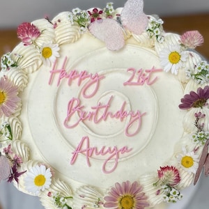 Peut inclure: Un gâteau rond et blanc glacé, décoré de fleurs roses, de papillons et du texte rose "Happy 21st Birthday Anya". Le gâteau est orné de marguerites et d'autres petites fleurs.