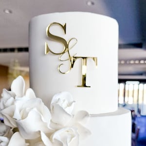 May include: A two-tiered white wedding cake decorated with white sugar roses and a gold mirror acrylic topper with the initials "S & T".