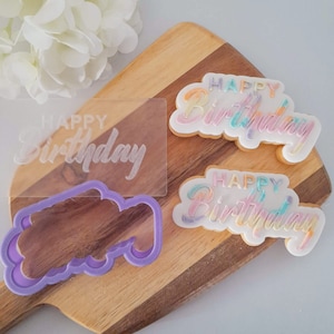 May include: A set of three cookie cutters and a stencil for making "Happy Birthday" cookies. The cutters and stencil are made of clear plastic and feature a rainbow-colored design.