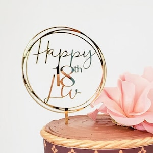 May include: A chocolate cake decorated with a brown and gold Louis Vuitton pattern, a pink rose, and a gold cake topper that says "Happy 18th Luv".