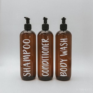 May include: Three amber-coloured plastic bottles with black pump dispensers. Each bottle features white vertical text labels reading SHAMPOO, CONDITIONER, and BODY WASH respectively. The bottles are cylindrical with a smooth, translucent finish.