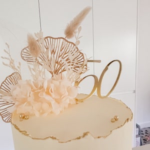 May include: A white cake with gold accents and a gold number 50 topper. The cake is decorated with dried flowers and leaves.