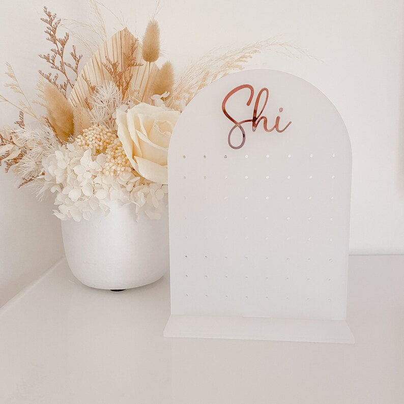 May include: A white acrylic earring display stand with an arched top and multiple holes for earrings. The word "Shi" is written in a copper-colored script on the top. A white vase with dried flowers and a rose is to the left.
