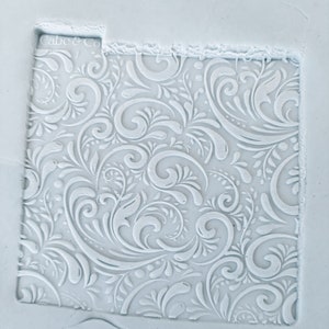 May include: White silicone mould with a raised design of a repeating pattern of white swirls and leaves. The mould is for making decorative fondant or cake toppers.