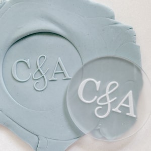 May include: A clear, round stamp with the letters "C&A" in white, used to create a wax seal. The stamp is placed on top of a light blue wax seal with the same letters.