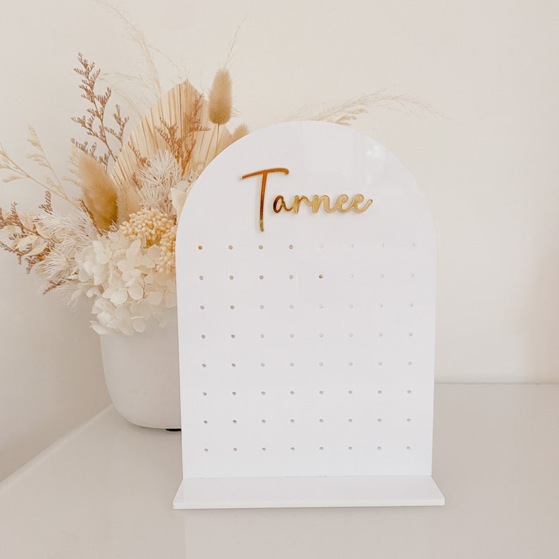 May include: A white acrylic earring display stand with an arched top and a series of small holes for hanging earrings. The name "Tarnee" is in gold script across the top. The stand is on a white surface.