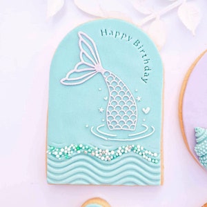 May include: A light blue sugar cookie decorated with a white mermaid tail and the words "Happy Birthday". The cookie has a wave pattern and white sprinkles around the edges.