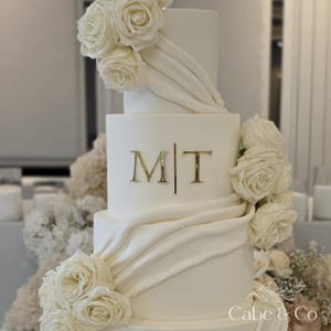 May include: A four-tiered white cake decorated with white roses and draped fabric. The cake features gold-coloured initials "M|T" and is adorned with floral arrangements. The bottom right corner of the cake has the text "Cabe & Co".