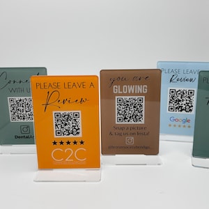 May include: Several acrylic review signs in different colours, each featuring a QR code and text like "Please Leave a Review" and "You are Glowing." The signs are displayed on clear stands.